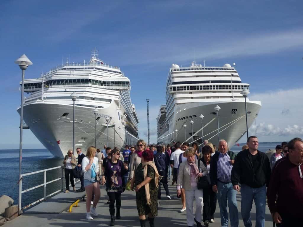 People walking on dock with two large cruise ships in background.