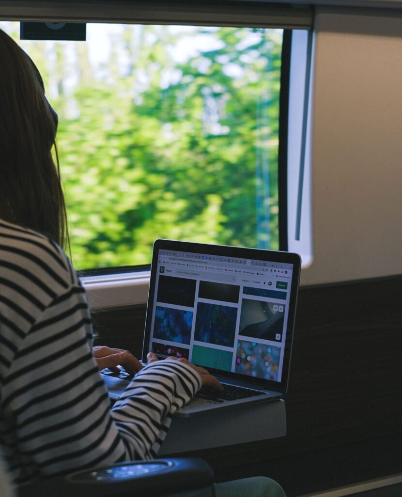 A woman working on a laptop inside a train, overlooking a scenic view.
